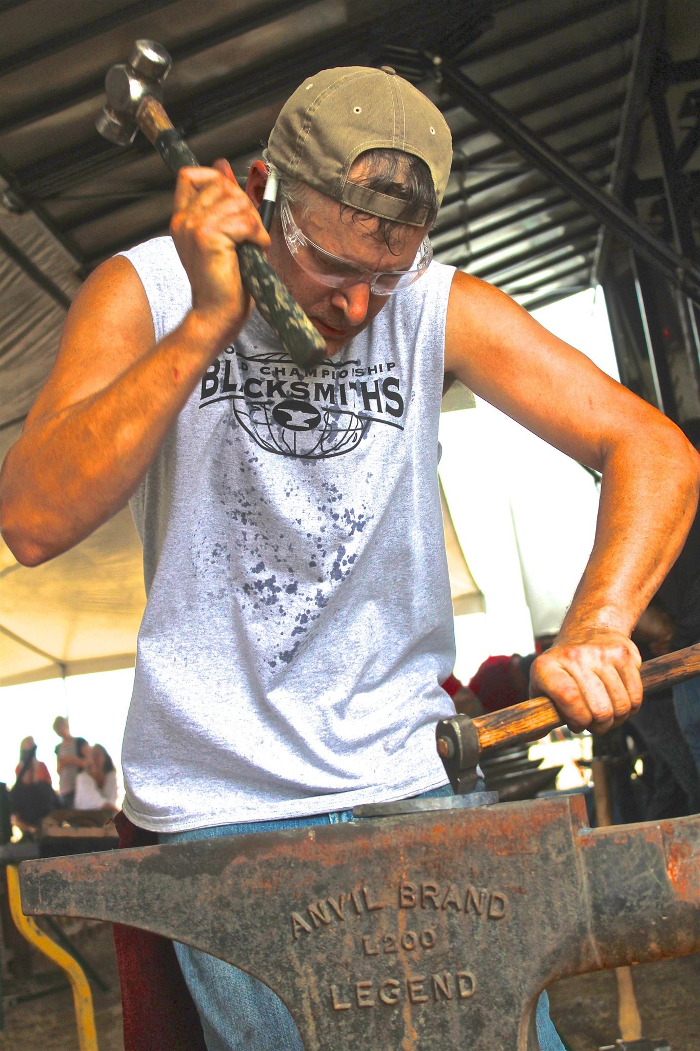 Steve wearing safety goggles and a cap hammers metal on an anvil, wearing a sleeveless shirt that reads 'Championship Blacksmiths.